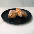 Two halves of a Cheesy Garlic Chicken Wrap, filled with chunks of chicken and sauce, are arranged on a round black plate atop a white marble surface with a light gray background.