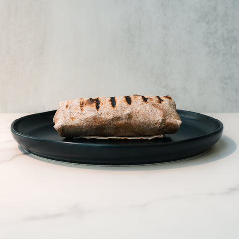 The Cheesy Garlic Chicken Wrap is served with visible char marks on a dark plate, set against a neutral background atop a light-colored surface.