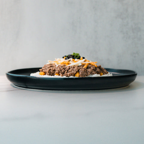 A black plate holds a Honey Garlic Beef Bowl with rice, ground beef, shredded cheese, and black olives on a light gray background.