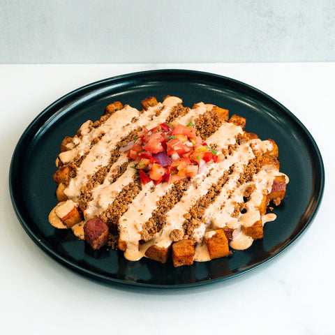 A black plate holds TexMex Potato Hash Subscription—a gluten-free dinner of crispy golden potato cubes with ground beef, creamy sauce, crumbled filling, and diced tomato, onion, and green garnish in the center.