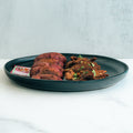 Garlic Flank Potatoes: Slices and shreds of cooked meat with herbs and sauce on a black plate—this high-protein meal is served on a white surface against a light background.