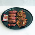 A black plate with sliced roasted purple sweet potatoes, spiced grilled meat garnished with herbs, and a packet of Sriracha hot sauce—an appetizing high-protein Garlic Flank Potatoes meal inspired by steak and potatoes.