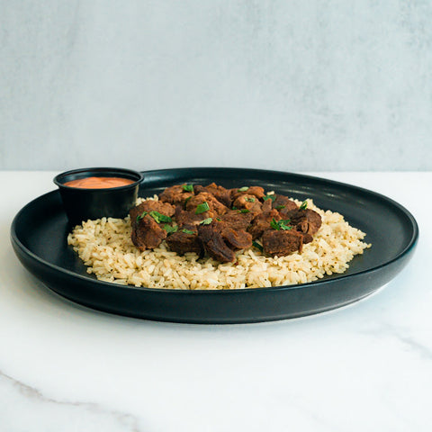 The GLORIOUS GAINS Steak Bites Cilantro Lime Rice features seasoned steak bites atop cilantro lime rice, garnished with herbs and served with a side of orange dipping sauce on a black plate, set on a white marble surface.