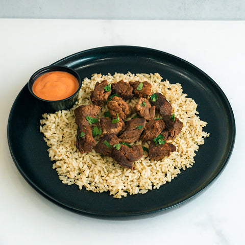 GLORIOUS GAINS Steak Bites Cilantro Lime Rice features cooked beef bites over cilantro lime rice, garnished with herbs and served with an orange dipping sauce—a perfect high-protein meal prep bowl.