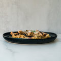 A black plate features Gluten Free Carbonara Chicken Subscription pasta, topped with grilled chicken and cheese, set on a light marble surface against a neutral background.