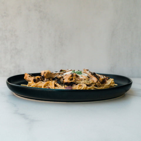 A black plate features Gluten Free Carbonara Chicken Subscription pasta, topped with grilled chicken and cheese, set on a light marble surface against a neutral background.