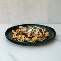 A black plate of Gluten Free Carbonara Chicken Subscription—high-protein pasta topped with grilled chicken, grated cheese, and herbs—sits on a white marble surface with a light textured background.
