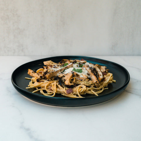 A black plate of Gluten Free Carbonara Chicken Subscription—high-protein pasta topped with grilled chicken, grated cheese, and herbs—sits on a white marble surface with a light textured background.