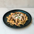 A black plate of Gluten Free Carbonara Chicken Subscription with high-protein pasta, grilled chicken, cheese, and herbs on white marble—a perfect Italian comfort food.