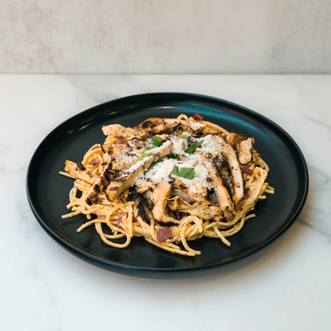 A black plate of Gluten Free Carbonara Chicken Subscription with high-protein pasta, grilled chicken, cheese, and herbs on white marble—a perfect Italian comfort food.