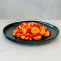 A black plate of Hawaiian Pineapple Shrimp 1lb, featuring gluten free shrimp and red bell peppers in a glossy sauce, sits on white marble with a light gray backdrop—a delectable, high-protein entrée.