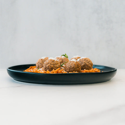 A black plate features Garden Orzo Meatballs topped with grated cheese, set on a white surface and light grey background—an ideal high-protein meal prep option.
