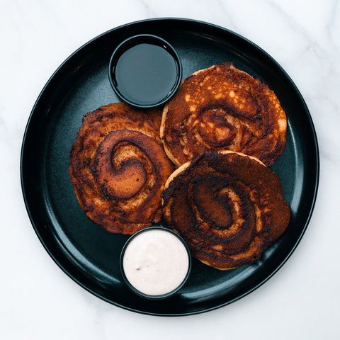Three Cinnamon Roll Pancakes are served on a black plate with a small cup of sugar-free syrup and cream sauce, set on a white marble surface.